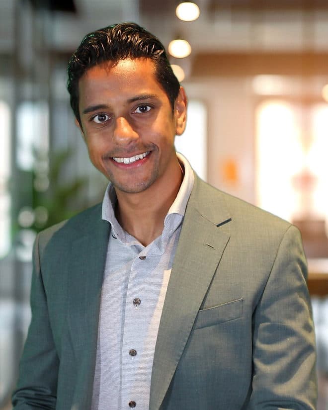 Ryan Audhoe, head and shoulders, in a grey-green jacket and an open light shirt, standing in an office with a glass wall behind him.