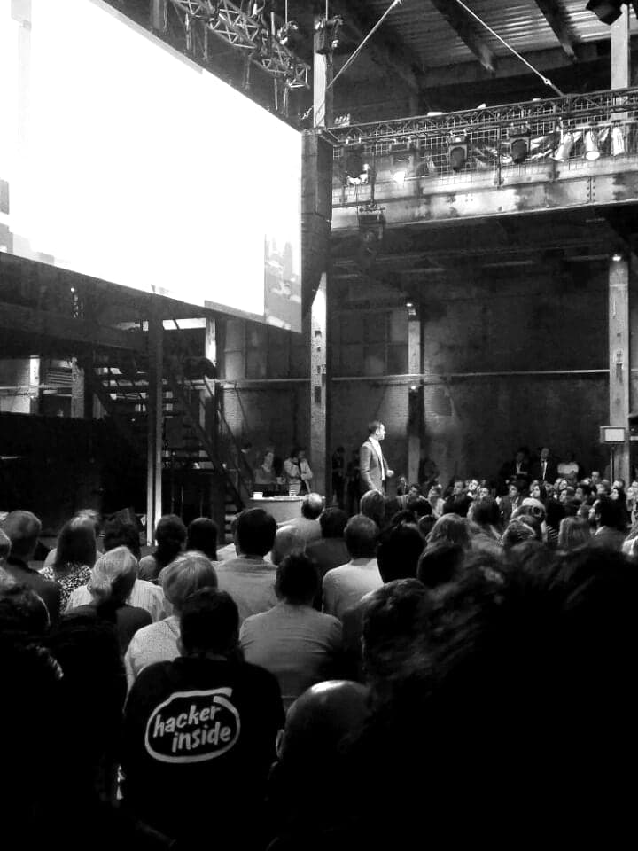 A talk in a converted industrial hall, seen from the back of a packed audience: a large projection screen above the stage, a single figure on stage, and rows of people standing and seated in front of it.